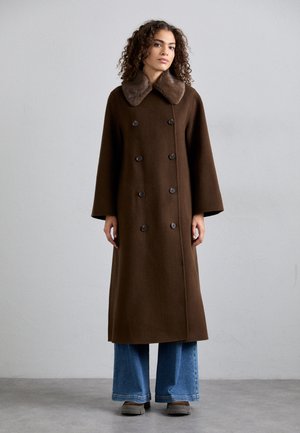 Brun uldfrakke med et faux fur-krave, dobbeltknapdesign og store knapper; har brede ærmer og en lang silhuet.