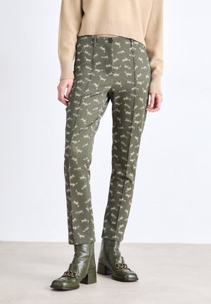 Olive green pants with a repeated tiger pattern in beige. Features side seams, a smooth texture, and a fitted design. Paired with green heeled boots.