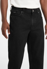 Melnas džinsa bikses ar taisnu griezumu, piecām kabatām, aizpogājamu rāvējslēdzēju un nelielu zīmola etiķeti priekšējā kabatā.