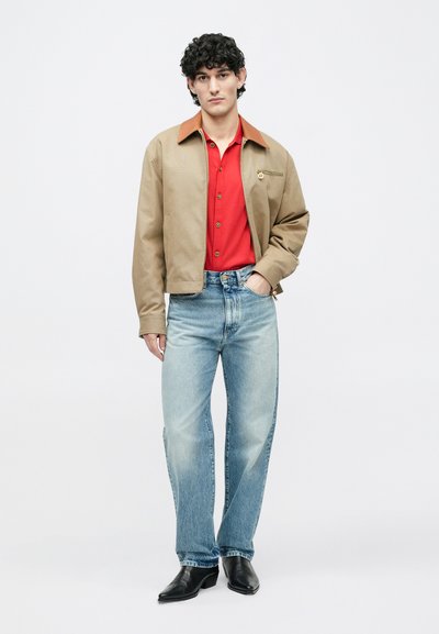 Käharate tumedate juustega mees, kes kannab beežit jopet punase särgi peal, helesiniseid teksaseid ja musti teravate tippudega saapaid, seisab ühe käega taskus.