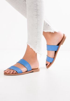 Niebieskie skórzane klapki z dwoma szerokimi paskami i płaską podeszwą, zestawione z jasnoszarymi dżinsami z postrzępionymi nogawkami.