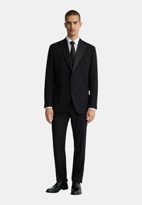 Completo nero con rever a punta, chiusura a un bottone, pantaloni coordinati e scarpe eleganti nere lucide. Camicia bianca e cravatta nera.