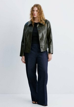 Mujer con cabello rizado rojo que lleva una chaqueta texturizada verde oscuro con botones, top negro, pantalones anchos azul marino y zapatos negros abiertos, de pie contra una pared blanca.