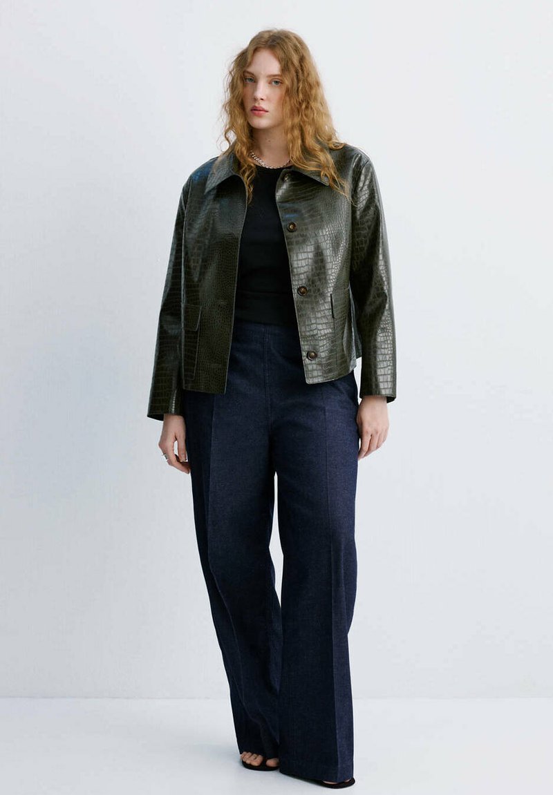 Femme aux cheveux roux bouclés portant une veste boutonnée texturée vert foncé, un haut noir, un pantalon large bleu marine et des chaussures noires à bout ouvert, debout contre un mur blanc.