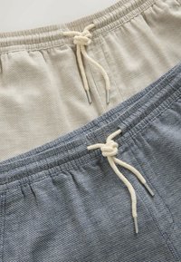 Shorts en tissu tricoté beige et bleu avec taille élastique et cordons de serrage crème noués, disposés en diagonale sur une surface claire.