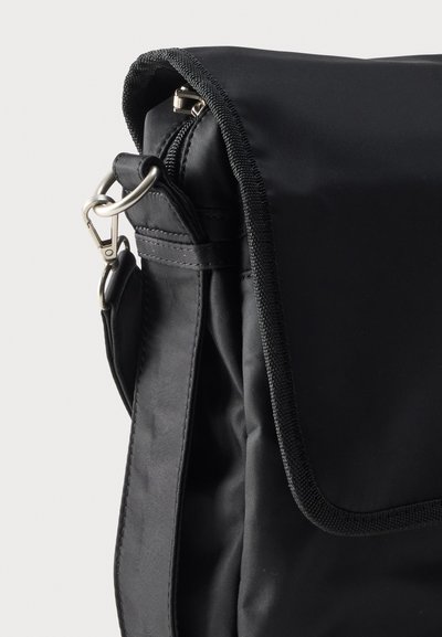 Μαύρη υφασμάτινη τσάντα ταχυδρόμου με μαξιλαρωμένο λουράκι, επάνω καπάκι και μεταλλικό κλιπ στη φωτεινή επιφάνεια.