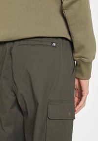 Olivgrön bomulls sweatshirt över mörkgrön cargobyxor, med en sidficka och en liten svart logotagg. Slät tygstruktur.