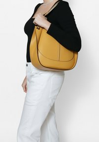 Geel leren handtas met een gebogen vorm, contrasterende donkere stiksels en een korte handgreep, gedragen over een zwarte top en witte broek.