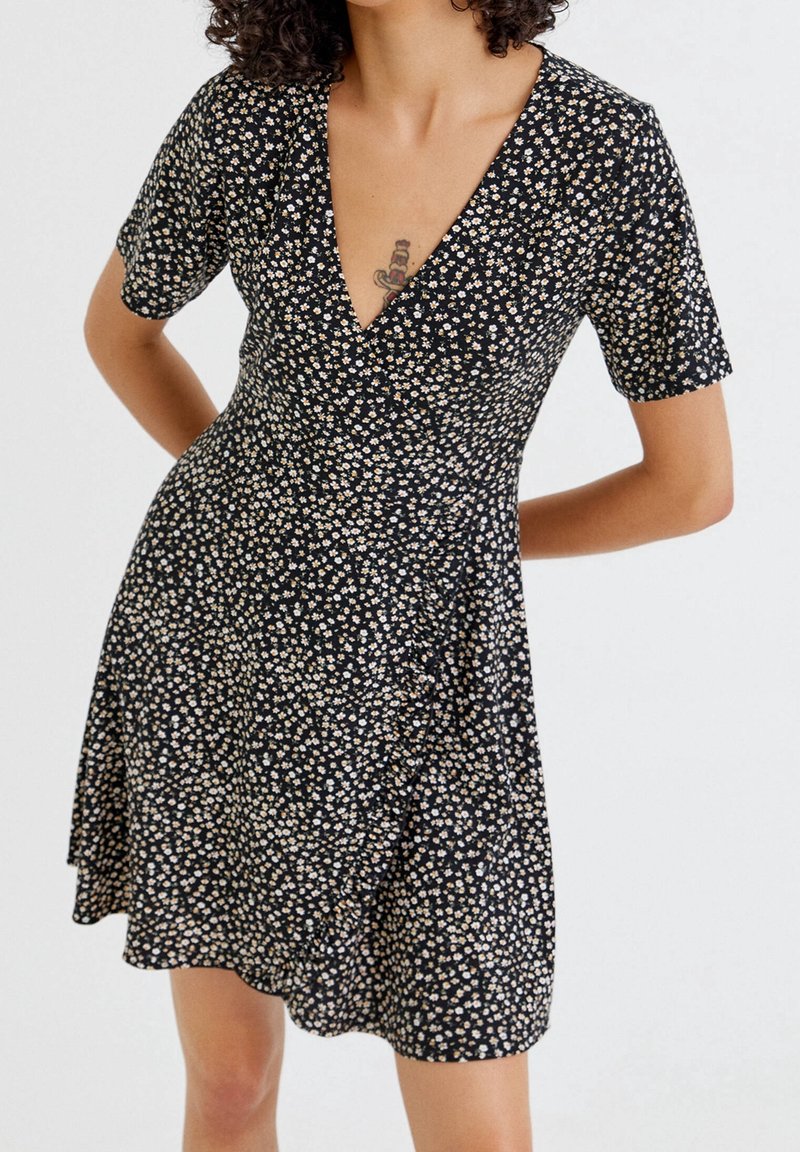 Robe noire avec un décolleté en V, des manches courtes et un motif floral en crème et beige, présentant un ourlet évasé et un léger ruché sur le côté.