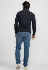 Pulover tricotat de culoare gri închis, cu guler înalt și tiv rezistat, purtat cu blugi din denim albastru deschis și adidași albi, văzut din spate.