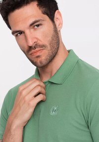 Zelená polo košile s límečkem, na hrudi s vyšitým emblémem. Tkanina vypadá texturovaně a prodyšně.