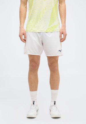 Mężczyzna w białych sportowych szortach i białych trampkach, stojący na tle jasnego, jednolitego tła, widoczny od talii do stóp.