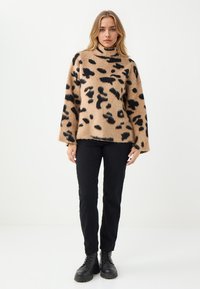 Beige trui met zwarte luipaardvlekken, gebreide textuur, hoge hals en brede mouwen. G gedragen met zwarte broeken en zwarte combatlaarzen.