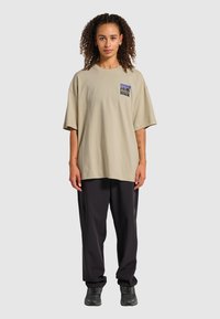Beige oversized t-shirt med en blocklogotyp på vänster bröst, kombinerad med lösa svarta byxor och mörka sneakers. Slät bomullstextur.