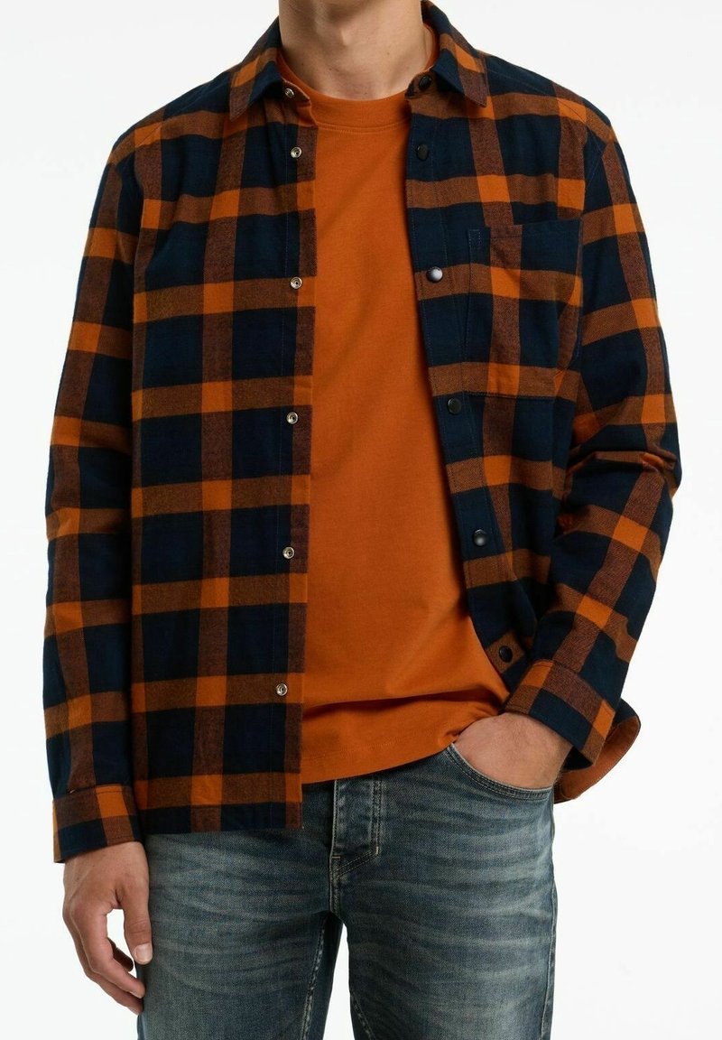 Personne portant une chemise à carreaux noire et orange déboutonnée sur un t-shirt orange et un jean bleu, avec une main dans la poche.