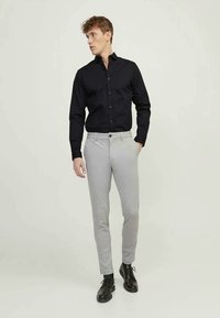 Homem vestindo uma camisa preta de botões, calças cinzas claras e sapatos pretos, de pé com uma mão no bolso, olhando de lado.