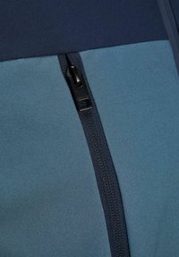 Sportos zakó, amely kék-zöld szövetből készült, sima tapintással és sötét cipzár részlettel az oldalon.