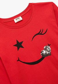Rød bomulds T-shirt med et sort smiley ansigt designet med en stjerne og et funkende hjerte lavet af palietter. Rund halsudskæring, korte ærmer.