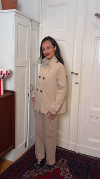 Kvinne med langt mørkt hår iført en beige dobbelspent frakk og bukser, stående innendørs nær hvite dører og et rødt mønstret teppe.