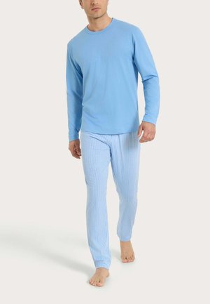 Uomo che indossa una camicia azzurro chiaro a maniche lunghe e pantaloni del pigiama a righe blu e bianche, che cammina a piedi nudi su uno sfondo bianco.