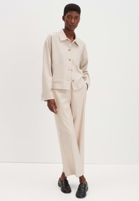 Beige twee-delige outfit met een blousejack met borstzakken en wijde broekspijpen, aangevuld met zwarte instappers.