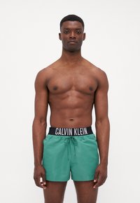 Zeleni kupaći šortsi s elastičnim pojasom i crnim Calvin Klein logotipom, s mogućnošću podešavanja uz pomoć vezice i glatkom teksturom.