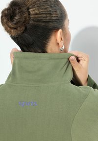 Olive green pullover with a high collar, featuring purple embroidery that reads "sports" at the back, made of soft, textured fabric.
