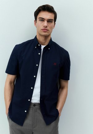 Navy short-sleeve button-up shirt with white buttons and an embroidered red logo, worn over a white t-shirt, paired with gray trousers.