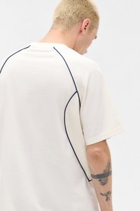 Cremefarvet t-shirt lavet af blød bomuld, har marineblå piping langs sømmene og et mesh-panel i siden for tekstur kontrast.