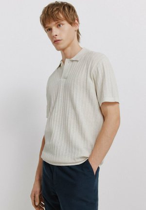 Młody mężczyzna o jasnobrązowych włosach, ubrany w kremową prążkowaną koszulkę polo z krótkim rękawem i ciemnoniebieskie spodnie, stoi z jedną ręką w kieszeni.