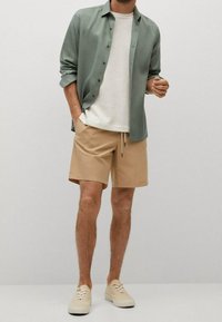 Lichtgroen overhemd over een crèmekleurig T-shirt, gecombineerd met beige shorts en lichte sneakers. Zachte textuur, casual ontwerp, ontspannen pasvorm.