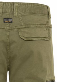 Olijfgroene shorts van stof met zichtbare stiksels. Voorzien van een patchzak aan de achterkant, een label met tekst en een rechte snit.