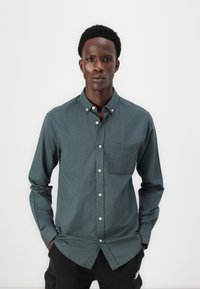 Camicia a maniche lunghe di colore verde scuro, con taschino sul petto, realizzata in cotone e dal design aderente. Presenta bottoni bianchi e una texture liscia.