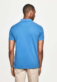 Modrá polokošile s krátkým rukávem a límečkem. Hladká bavlněná látka, jemné švy, nošena s béžovými kalhotami. Pohled zezadu.