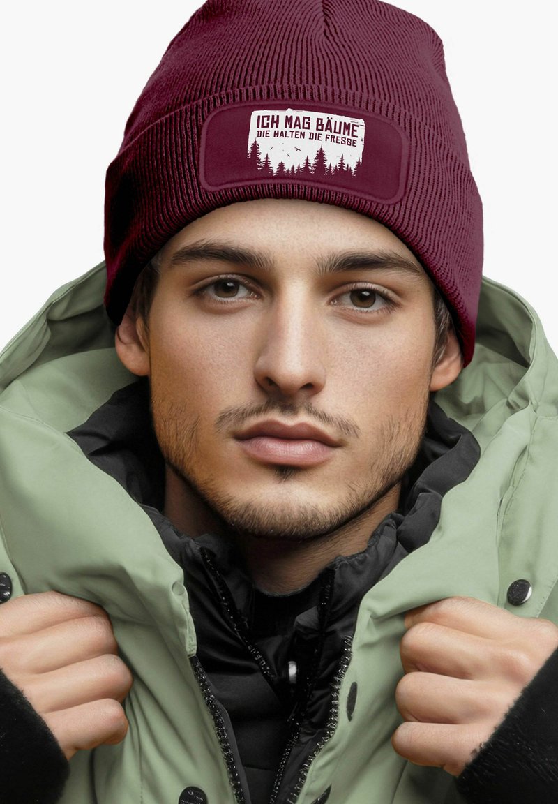 Burgundy knit beanie with a forest-themed patch; wearing a light green coat over a black zip-up. Texture is ribbed and smooth.