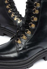 Botas de cuero negro con ojales dorados, suela negra gruesa y sistema de cordones prominente. Textura suave y diseño de puntera reforzada.