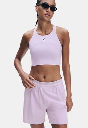 Donna che indossa un crop top sportivo lilla chiaro e pantaloncini con occhiali da sole neri, posando con una mano sui fianchi contro uno sfondo uniforme.