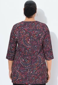 Vrouw met opgestoken haar, die grote ringoorbellen draagt en een tuniek met lange mouwen in een rood en zwart paisleypatroon, gezien vanuit de achterkant.