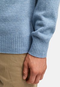 Lyseblå strikket genser med ribbestrikket nederkant og mansjetter, laget av mykt materiale, sammen med lysebeige stoffbukser, med synlige hender hvilende på stoffet.