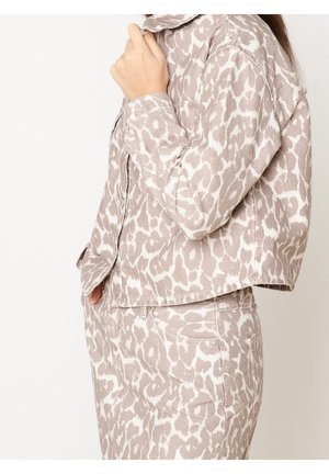 Femme portant une veste et un pantalon assortis beige et blanc à motif léopard, debout avec une main près du col et l'autre dans la poche.