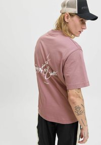 Rosa oversized t-shirt med korte ærmer, der har hvid grafisk tekst på ryggen. Båret med sorte bukser og en sort kasket.