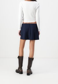 Top a maniche lunghe bianco a costine abbinato a una gonna plissettata blu navy e stivali marroni fino al ginocchio con fibbie e una texture liscia.