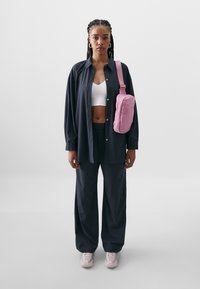 Svart oversized knappskjorte, hvit kort topp, marineblå vid bukse, rosa crossover-veske og lyserosa sneakers. Nøytral bakgrunn.