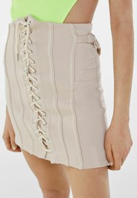 Jupe mini beige avec des coutures verticales, un devant à lacets avec œillets en argent et de larges lacets, et des poches latérales avec des détails de couture.