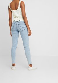 Vaaleanvihreä hihaton toppi, jossa teksturoitu kuvio ja röyhelöinen helma, yhdistettynä vaaleansinisiin kapeisiin farkkuihin ja valkoisiin tennareihin, nähtynä takaa.