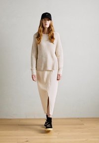 Femeie cu păr lung, îmbrăcată cu un pulover bej cu dungi, fustă asortată cu despicătură în față, șapcă neagră, șosete negre și adidași negri, mergând în interior.