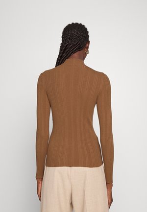 Vrouw met donker gevlochten haar, gouden oorringen in de vorm van hoepels, een nauwsluitend bruin geribd shirt met lange mouwen en lichtbeige broek, gezicht naar achteren gekeerd.