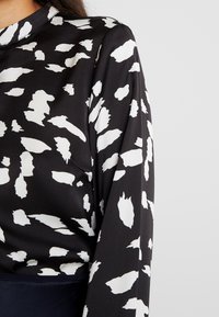 Blouse noire avec motif abstrait blanc, manches longues, tissu doux, col rond et coupe légèrement ample. Pas de quincaillerie ou d'accents visibles.