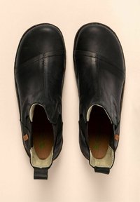 Un par de zapatos negros de piel tipo slip-on con punteras redondas y paneles laterales elásticos, vistos desde arriba sobre un fondo beige.
