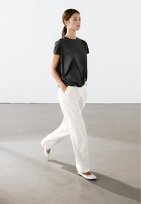 Sort kortærmet skjorte, løse hvide pinstribede bukser og hvide flade sko. Model går på en neutral gulv med en lys baggrund.